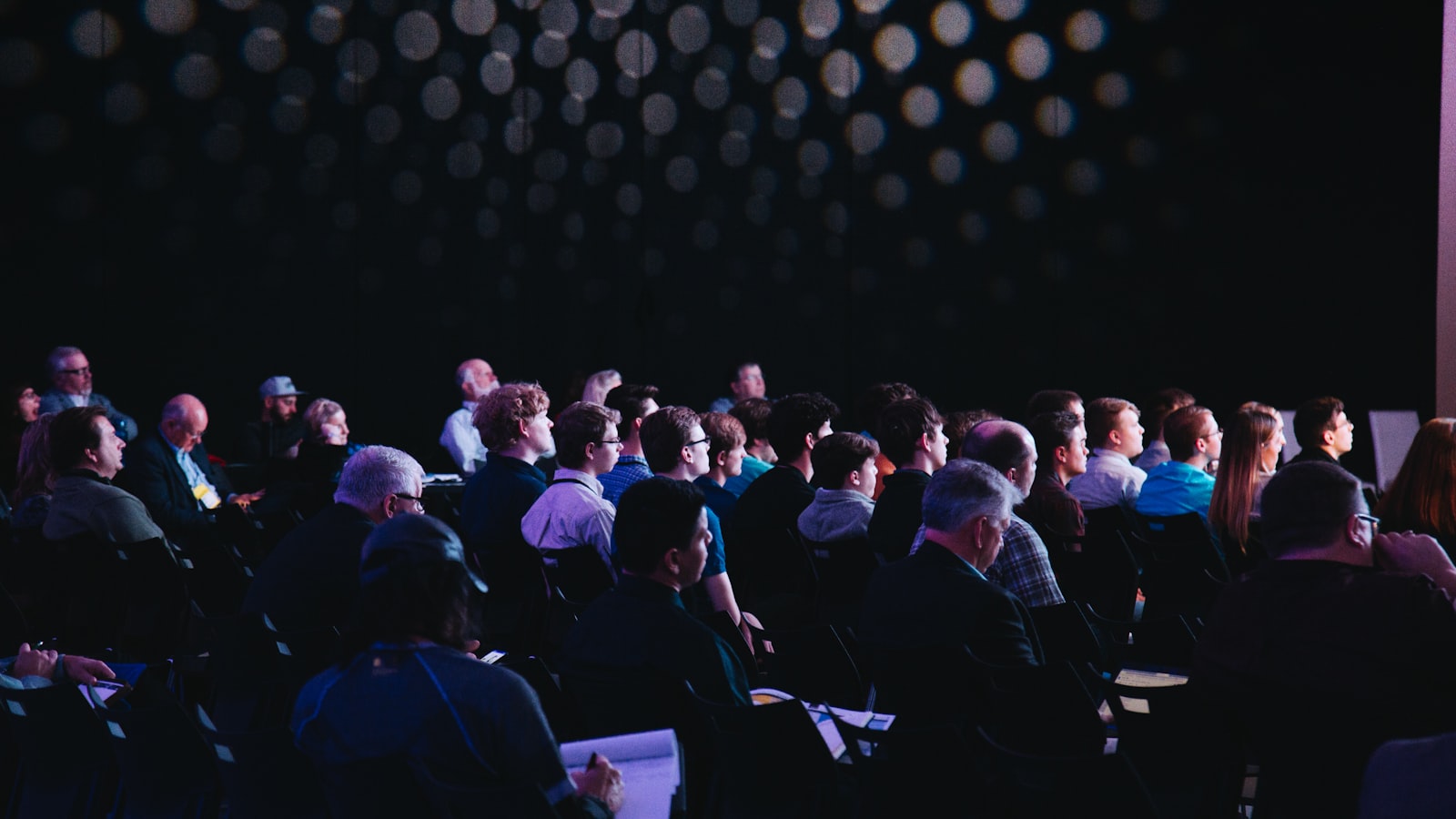 Audience seated in a darkened conference hall, facing a brightly lit stage