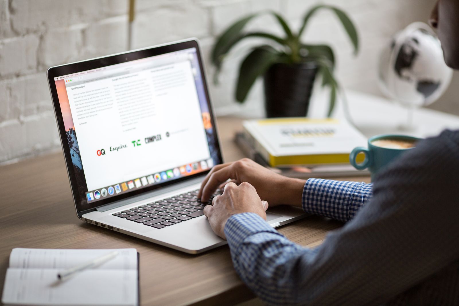 Person in a checked shirt typing on a silver laptop at a light wooden home desk