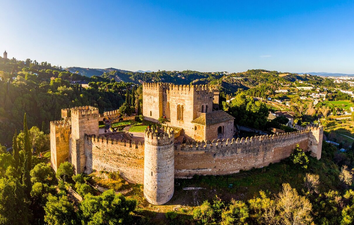 Aerial view of a fortified stone castle with crenellated walls and towers on a green hilltop at sunset, forested hillsides and distant mountains behind