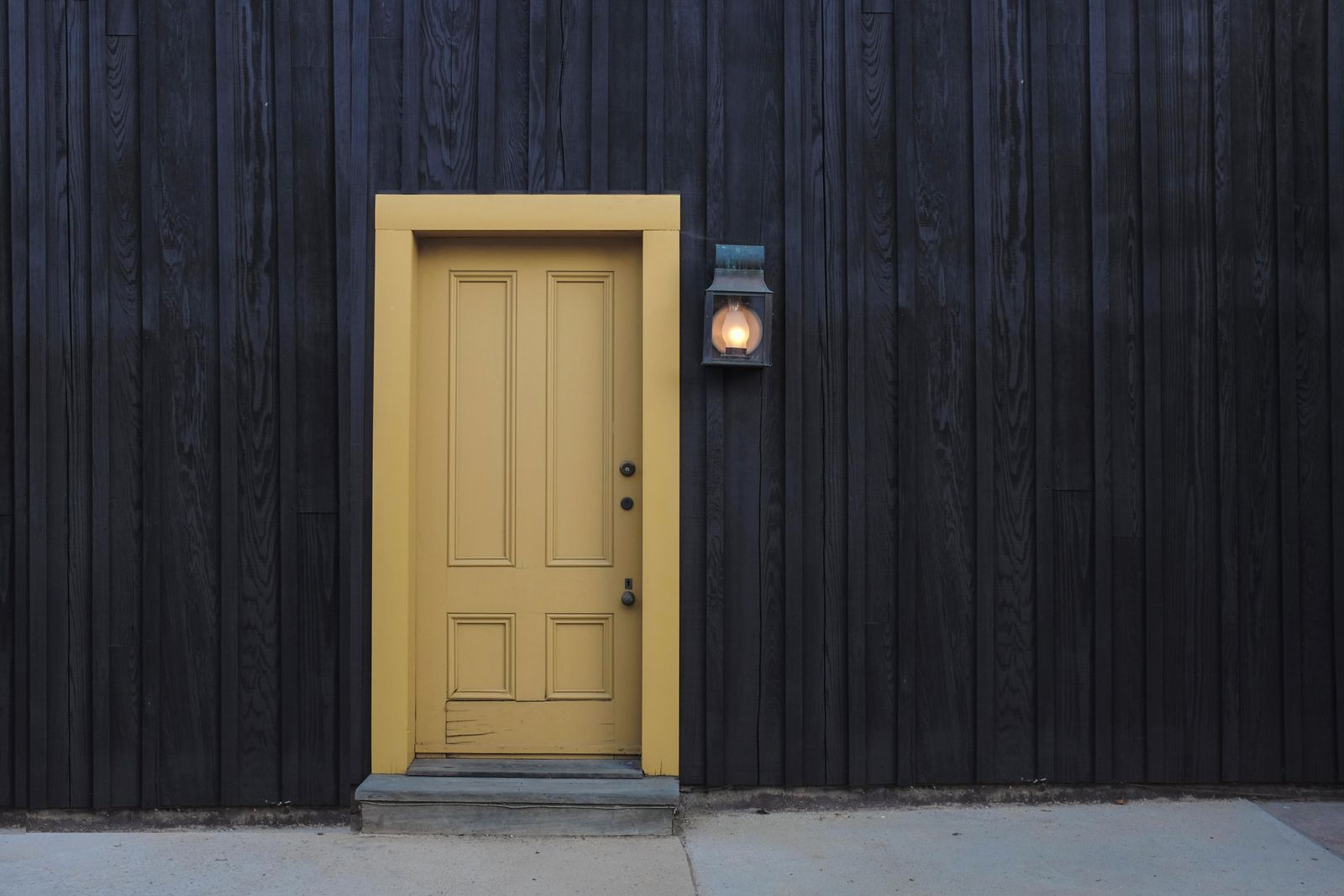 Single pale-yellow door set in a black wooden wall, with a small lantern beside the frame