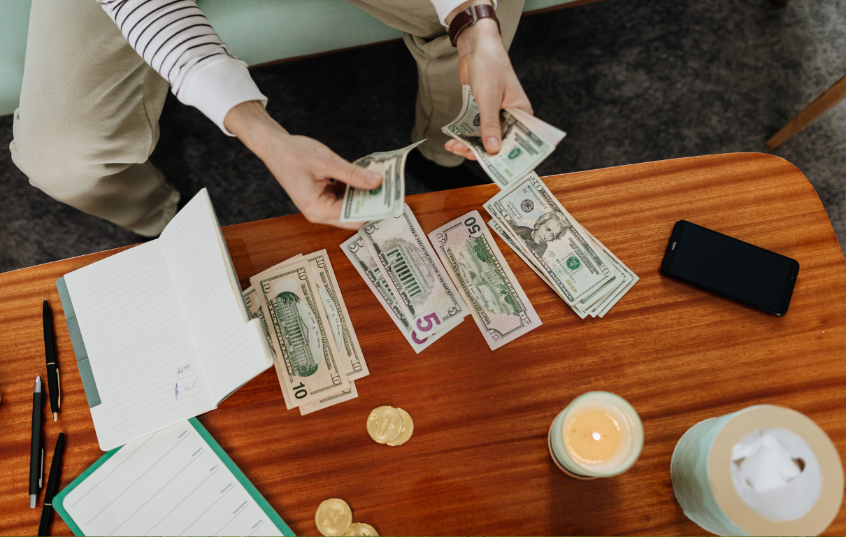 Hands counting cash bills at a wooden table beside a notebook and coffee — the daily morning finance check ritual