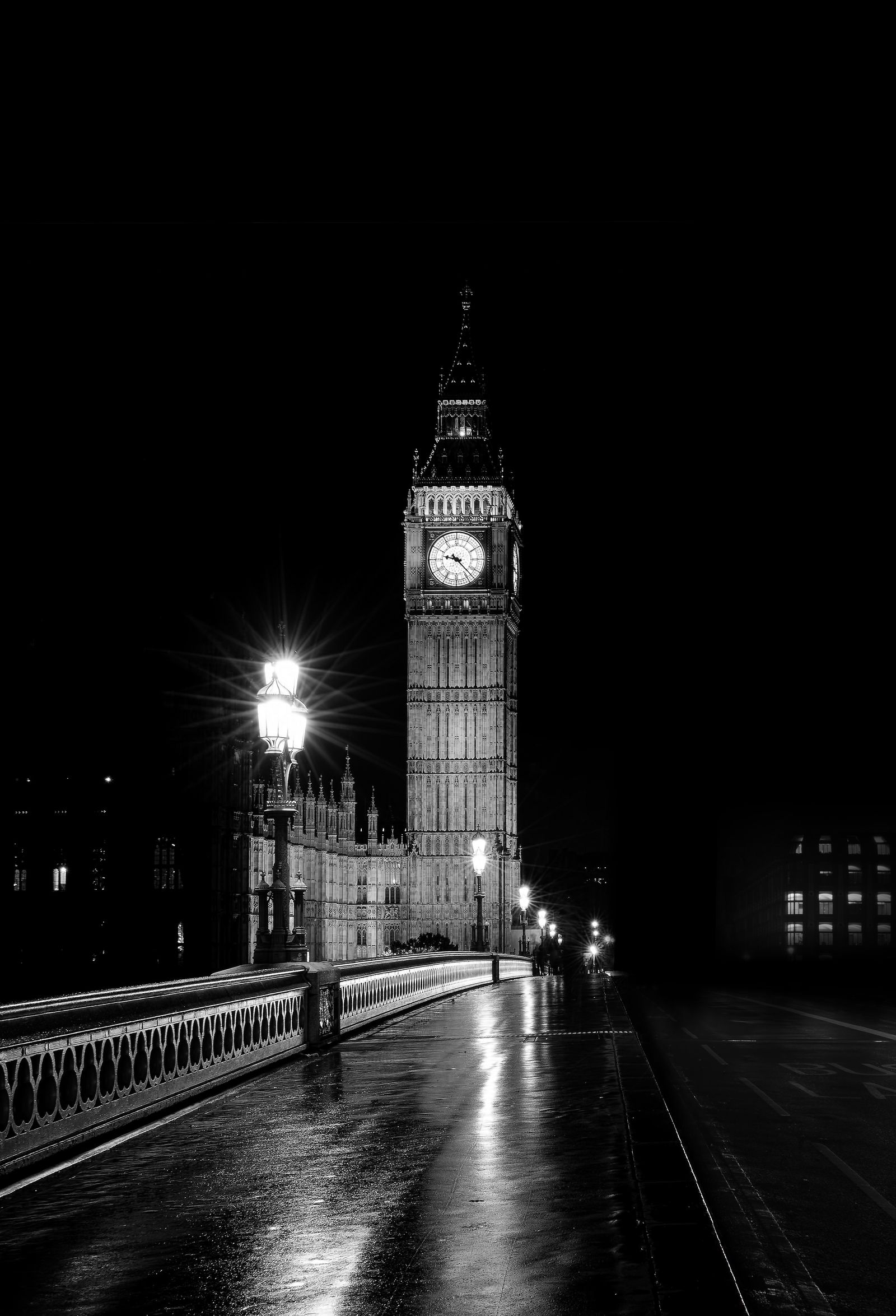 Clock tower at night above a wet street and glowing lamps, photographed in black and white